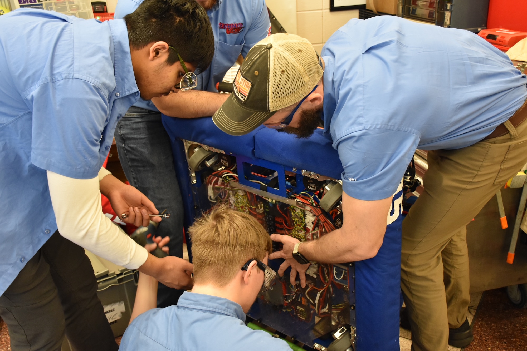 A man and two kids, all in light blue, short sleeve shirts and safety goggles, work on a robot. Multiple wires are visible.