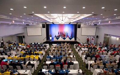 A photo of a large room filled with people facing a person speaking on stage at a podium.