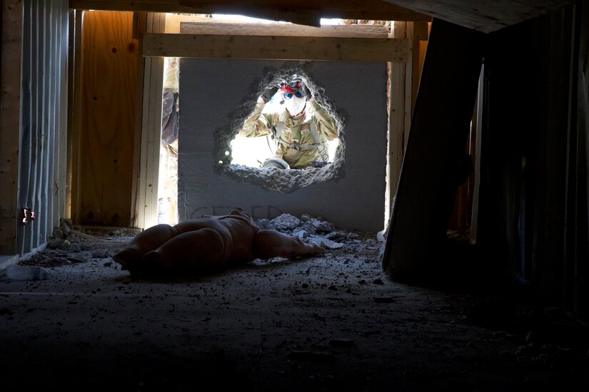 A service member looks through a hole that has been cut in concrete to reveal a simulated casualty on the other side.
