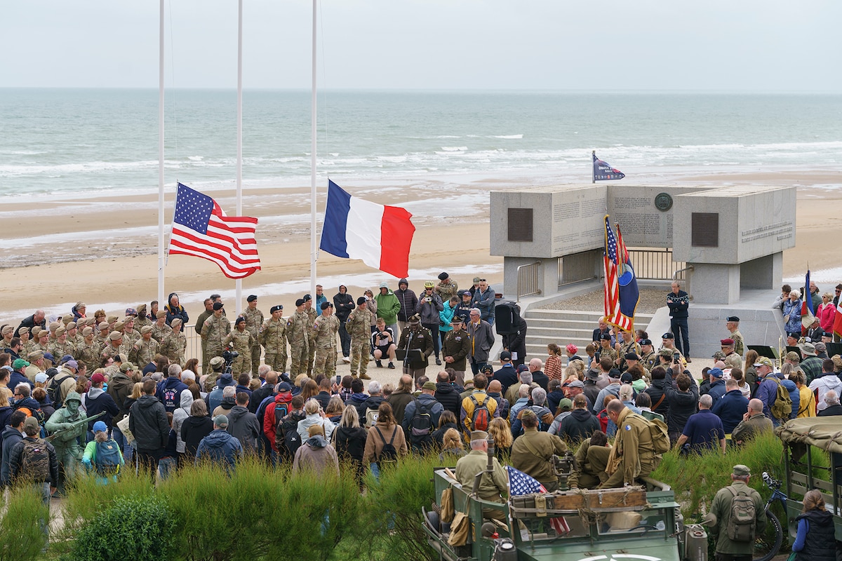 A crowd gathers at a monument on a beach where U.S. and French flags fly at half staff.