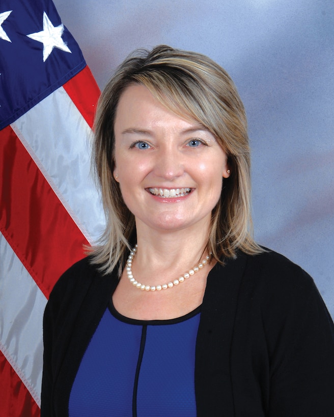 Fair skinned woman with blond shoulder length hair stands in front of a U.S. Flag and is wearing a blue shirt, black cardigan sweater and a pearl necklace.