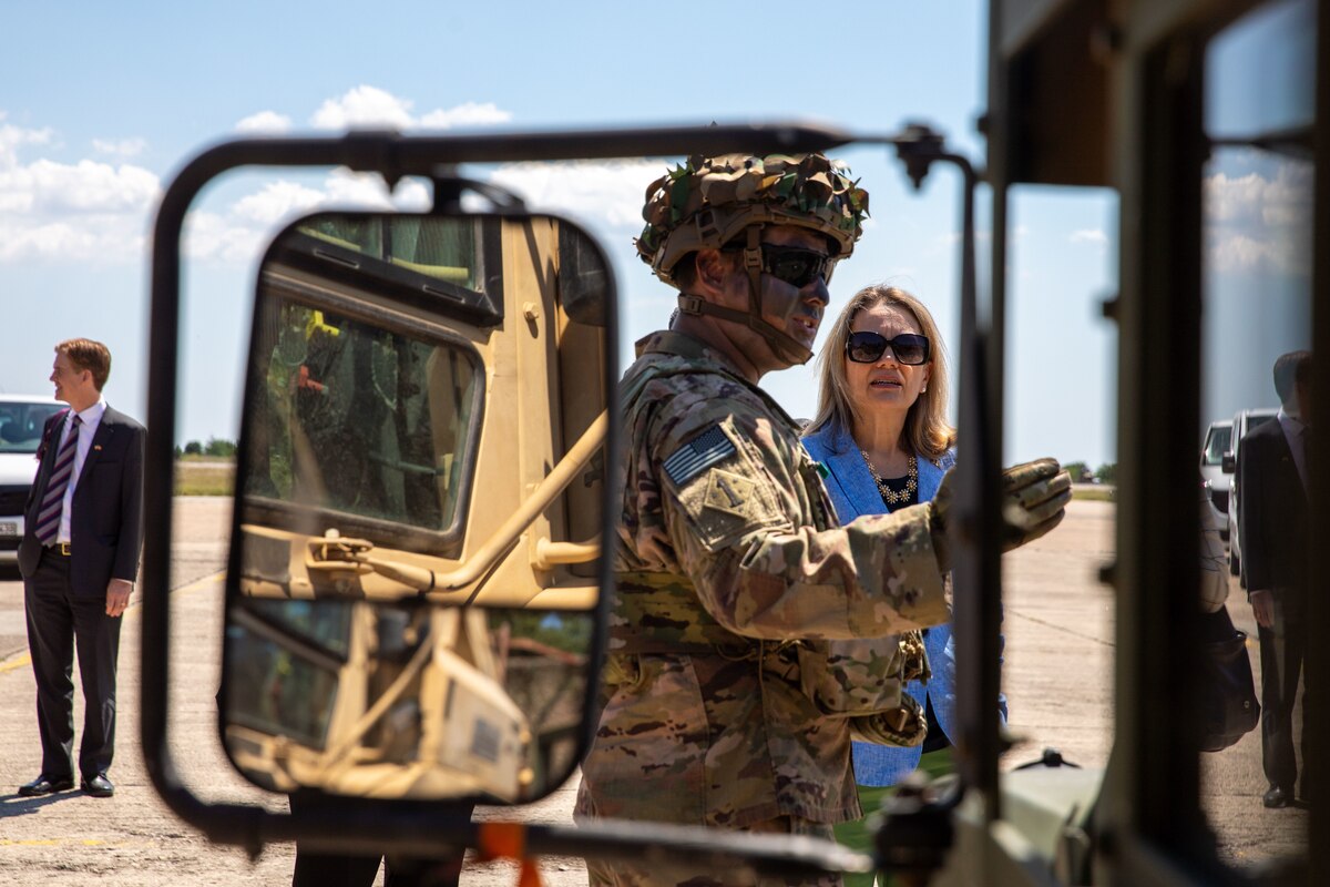 A man wearing camouflage uniform motions toward something off camera as a woman looks on.