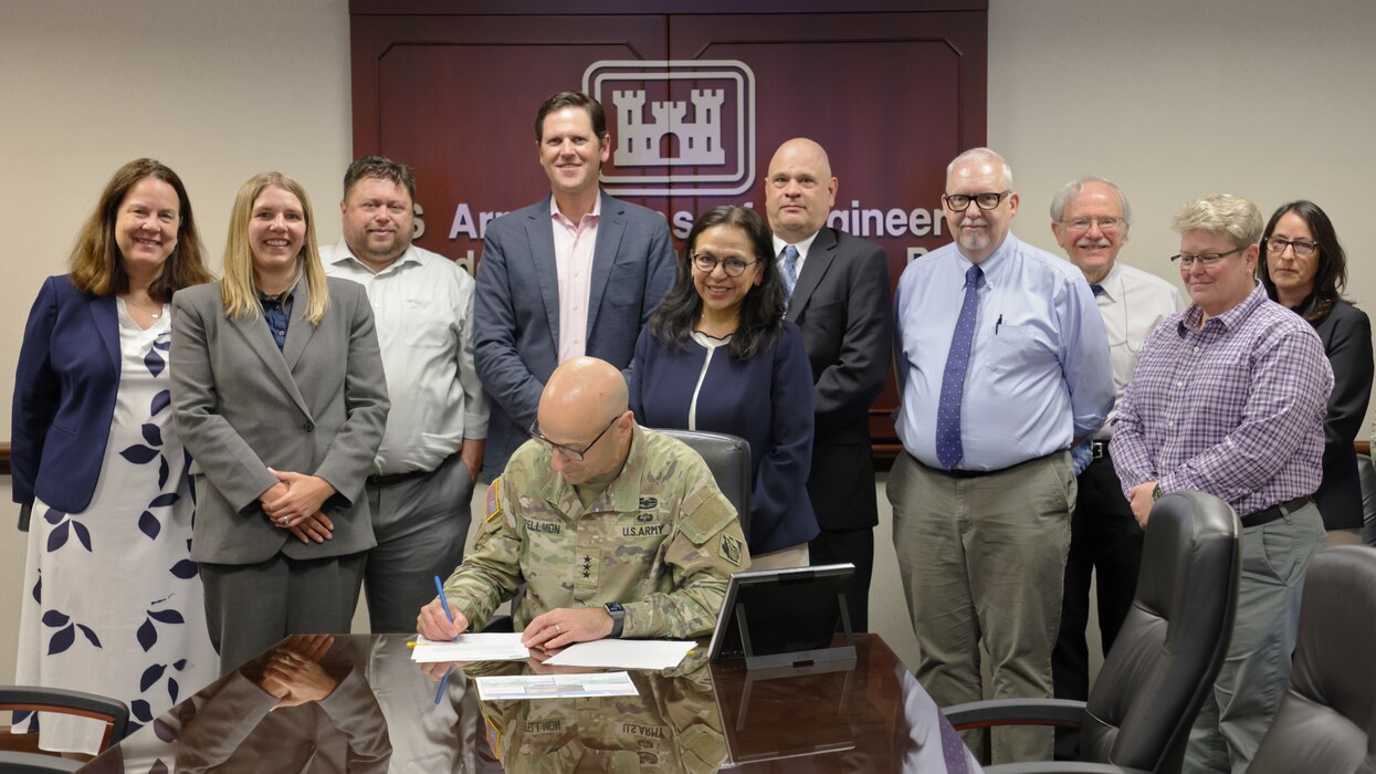 10 white men and women stand behind a seated white male soldier in green camo as he signs a document on a brown table.