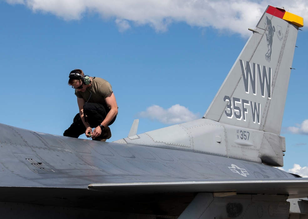A maintainer stands on top of an F-16 aircraft lifting up a panel.