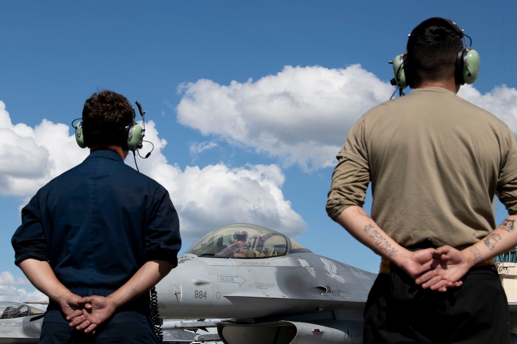 Two maintainers stand side by side with their hands behind their back waiting to marshal an F-16 aircraft out for takeoff.