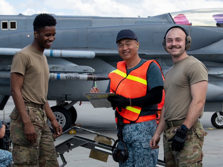 Two U.S. Air force members pose with a Republic of Singapore Air Force member for a group photo on the flightline in front of an aircraft.