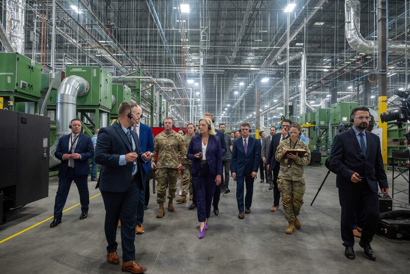 Service members and civilians walk in a group through a large room in an industrial facility.
