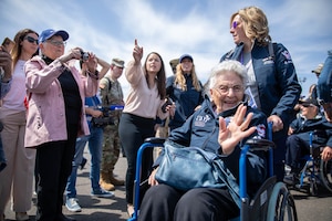 A veteran at the front of a procession of fellow vets smiles and waves as a crowd of people take photos and watch on a flight line.