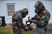 Two soldiers wearing full protective gear adjust settings and learn to use the chemical vapor detector.