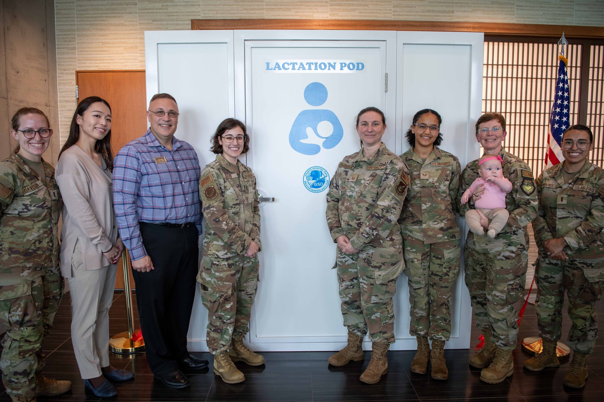 a group of military members and civilians gather infront of a lactation pod for a photo