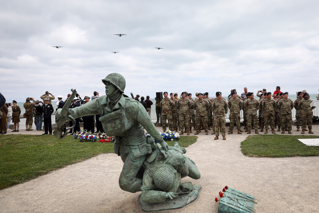 A large group of service members stand facing a beachside statue of a service member pulling a wounded comrade as four aircraft fly in formation overhead.