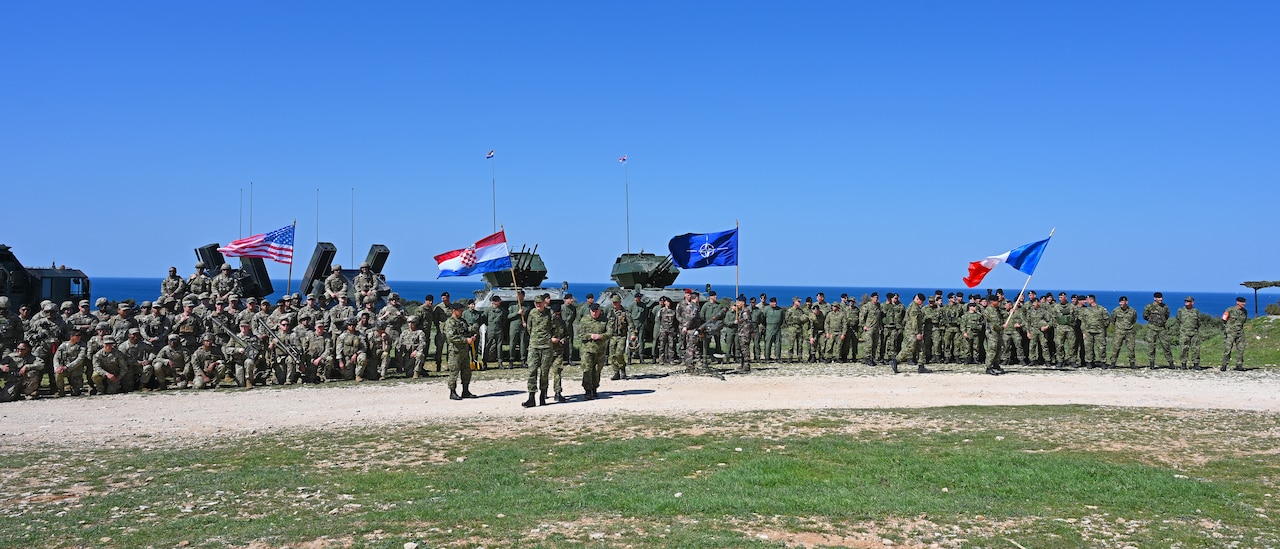 A large number of service members fly four different flags as they pose for a photo on a clear day.