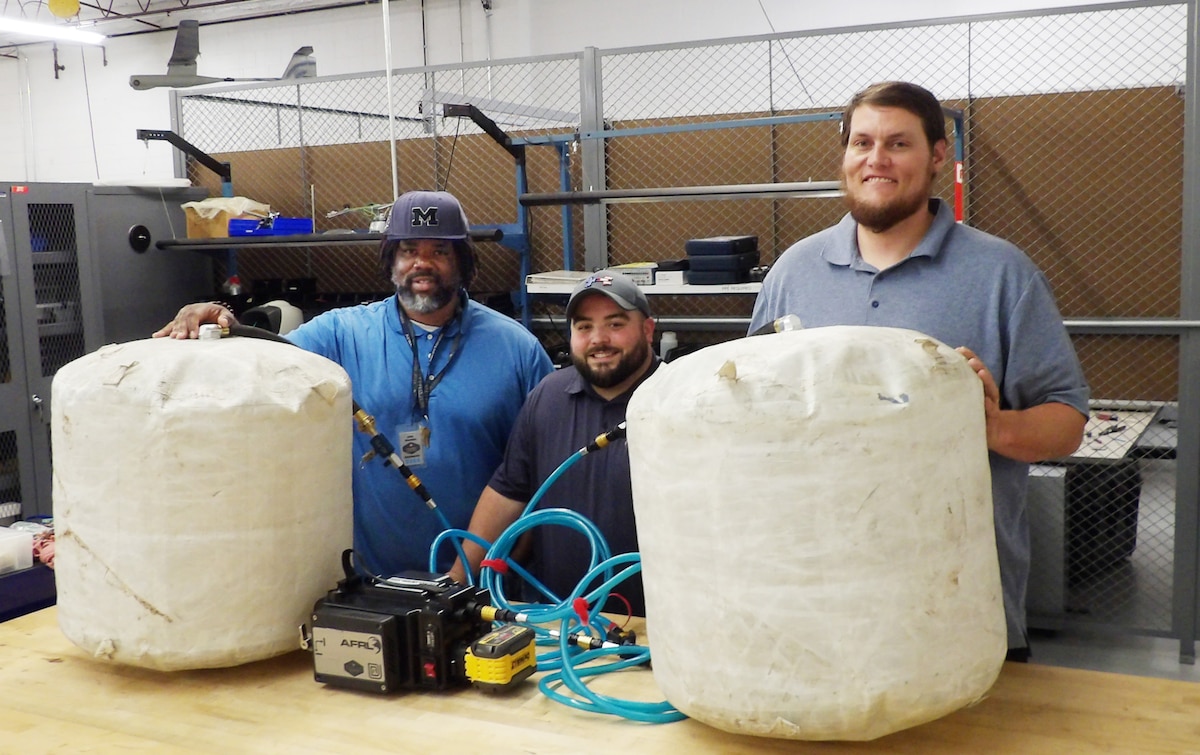 Three engineers stand with the air compressor and two large bags of the Heavy Lift Kit, which first responders can use to lift heavy objects.