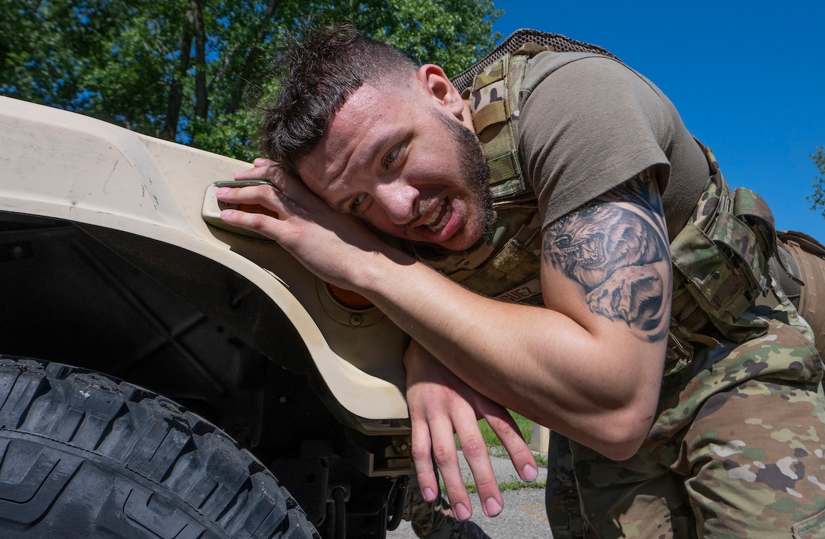 A muscular, tattooed Airman pushes with his shoulder against the back of a vehicle.