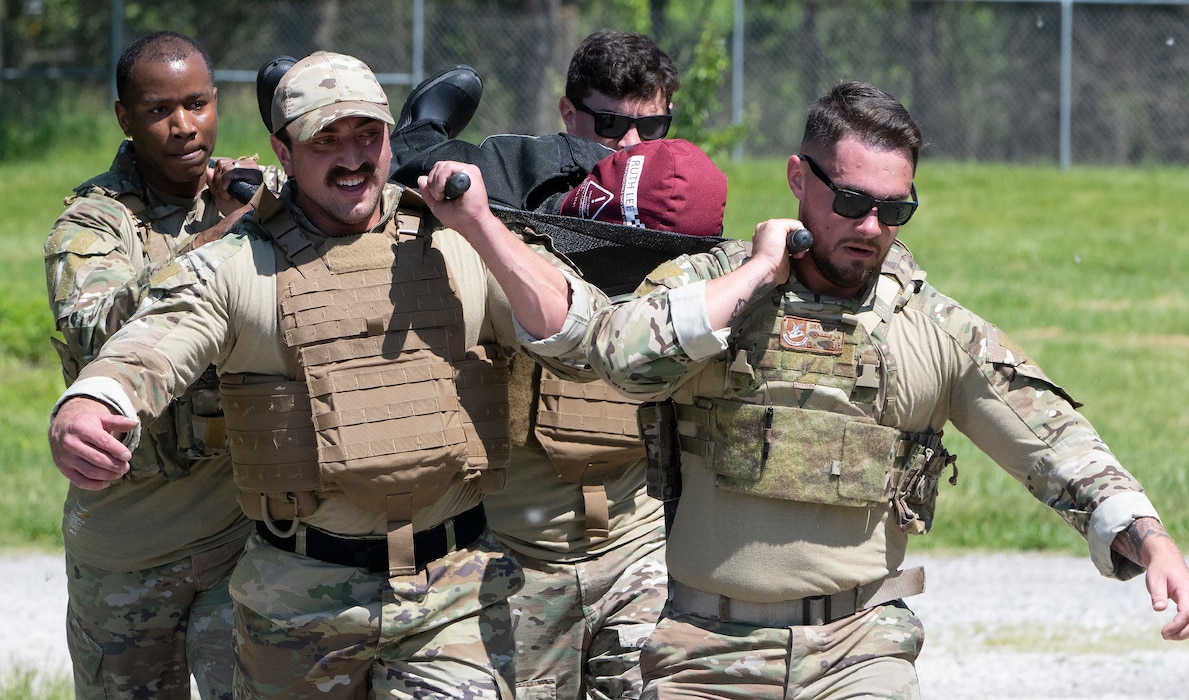 Four men in camouflage uniforms carry a stretcher, with a mannequin, on their shoulders.