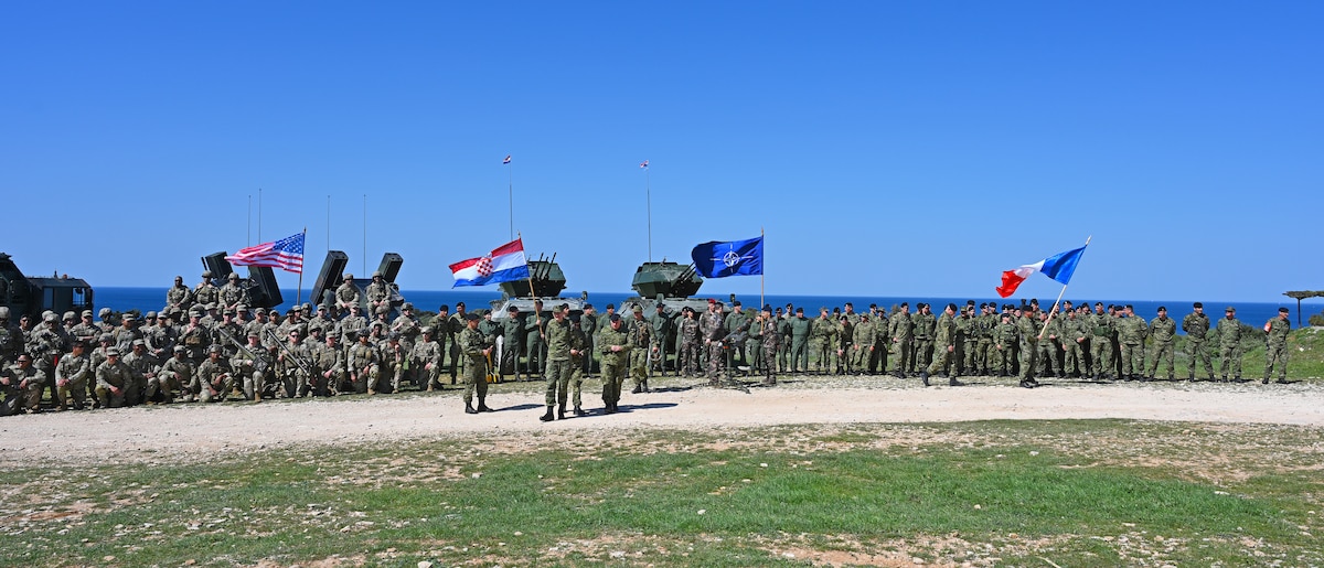 A large number of service members fly four different flags as they pose for a photo on a clear day.