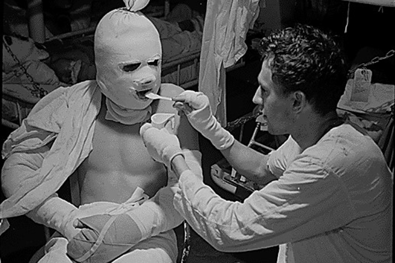 A person feeds another whose head and arms are covered in bandages.