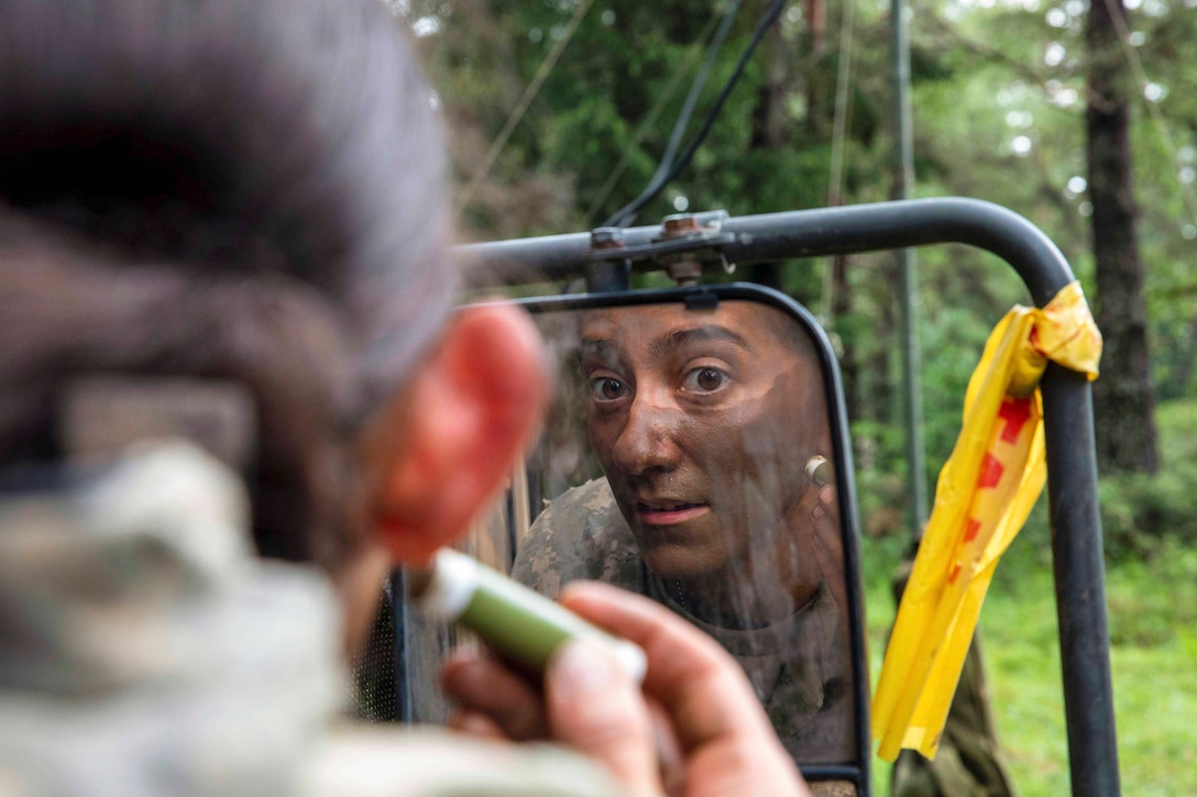 A soldier uses a makeup stick to put camouflage on their face while looking in a vehicle’s sideview mirror.