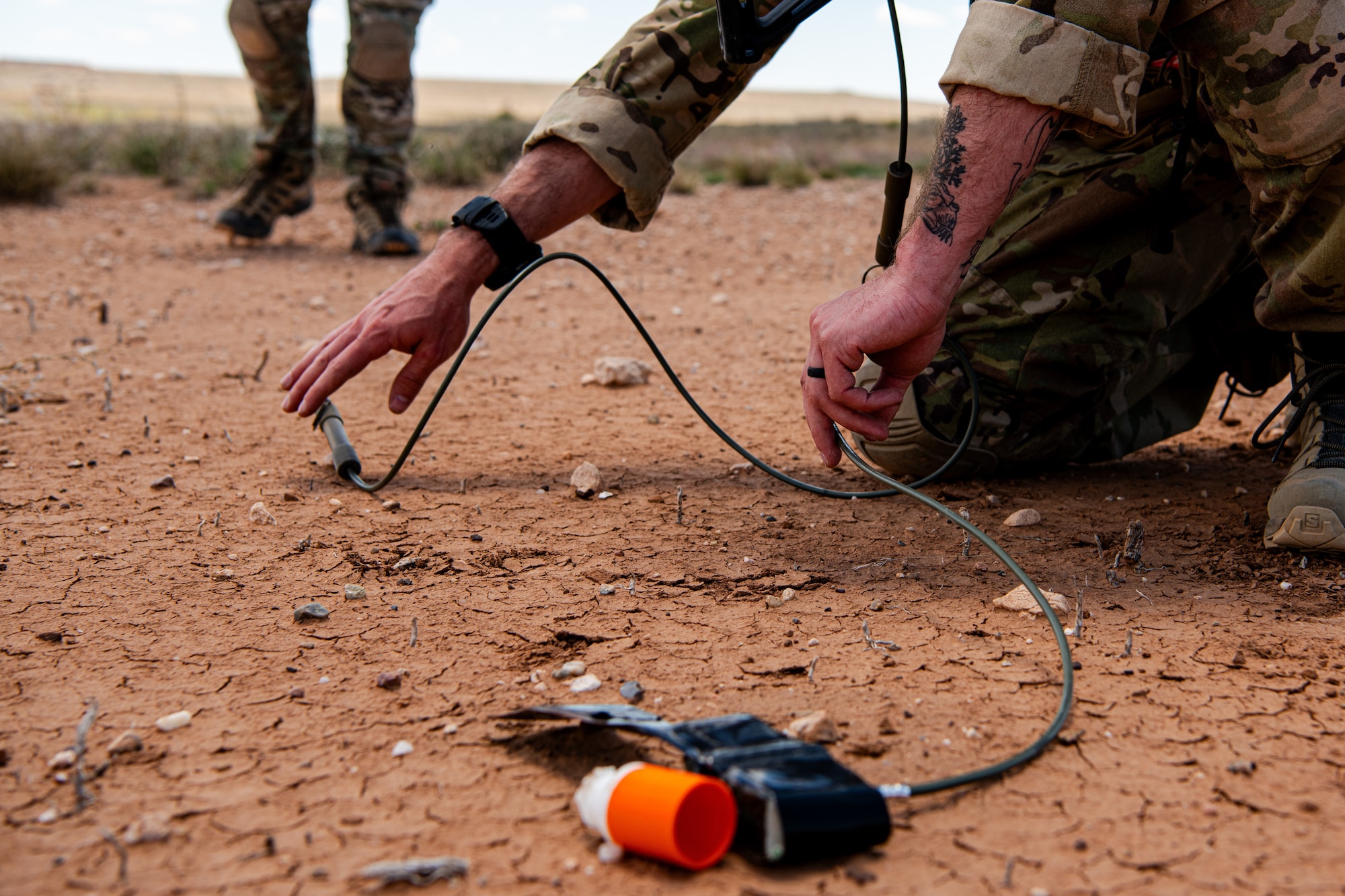 A man in military uniform kneels and reaches for military demolition material on the ground.