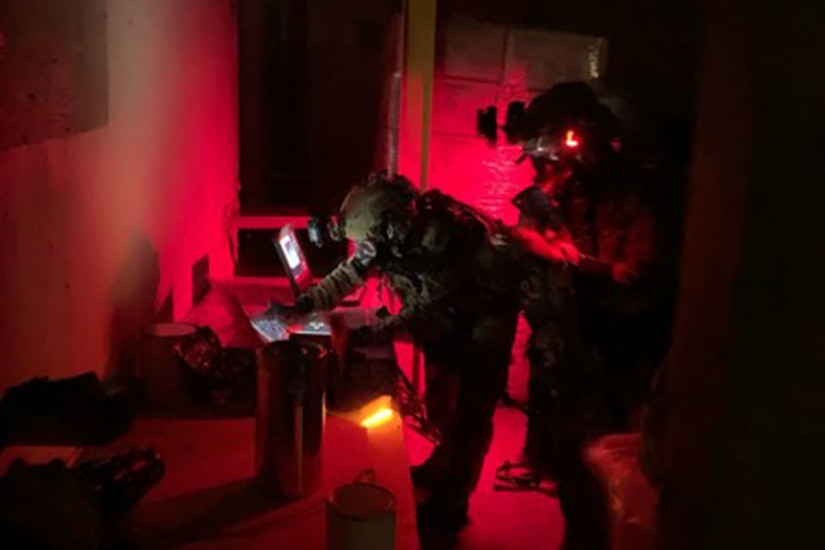 Two service members work on equipment in a darkened room with red light reflected.