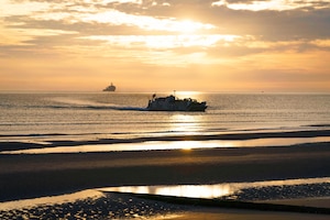 A boat approaches the shore as a ship sails in the distance under a sunlit sky.