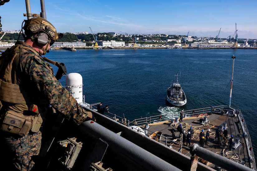 A Marine stands watch aboard a ship while looking down a sailors working on a lower deck.