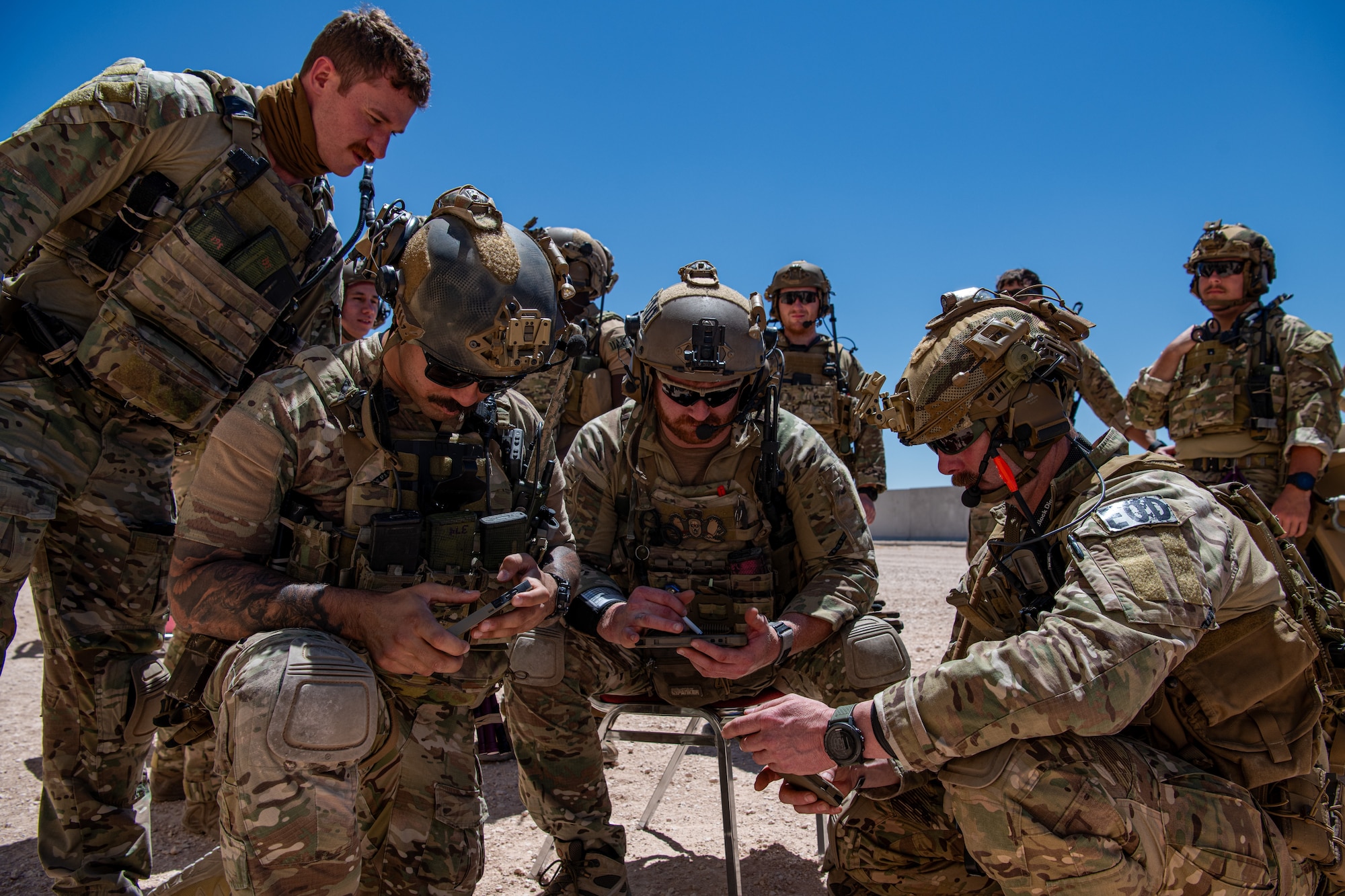 A group of men in military uniform and military equipment stand and kneel in a circle planning.