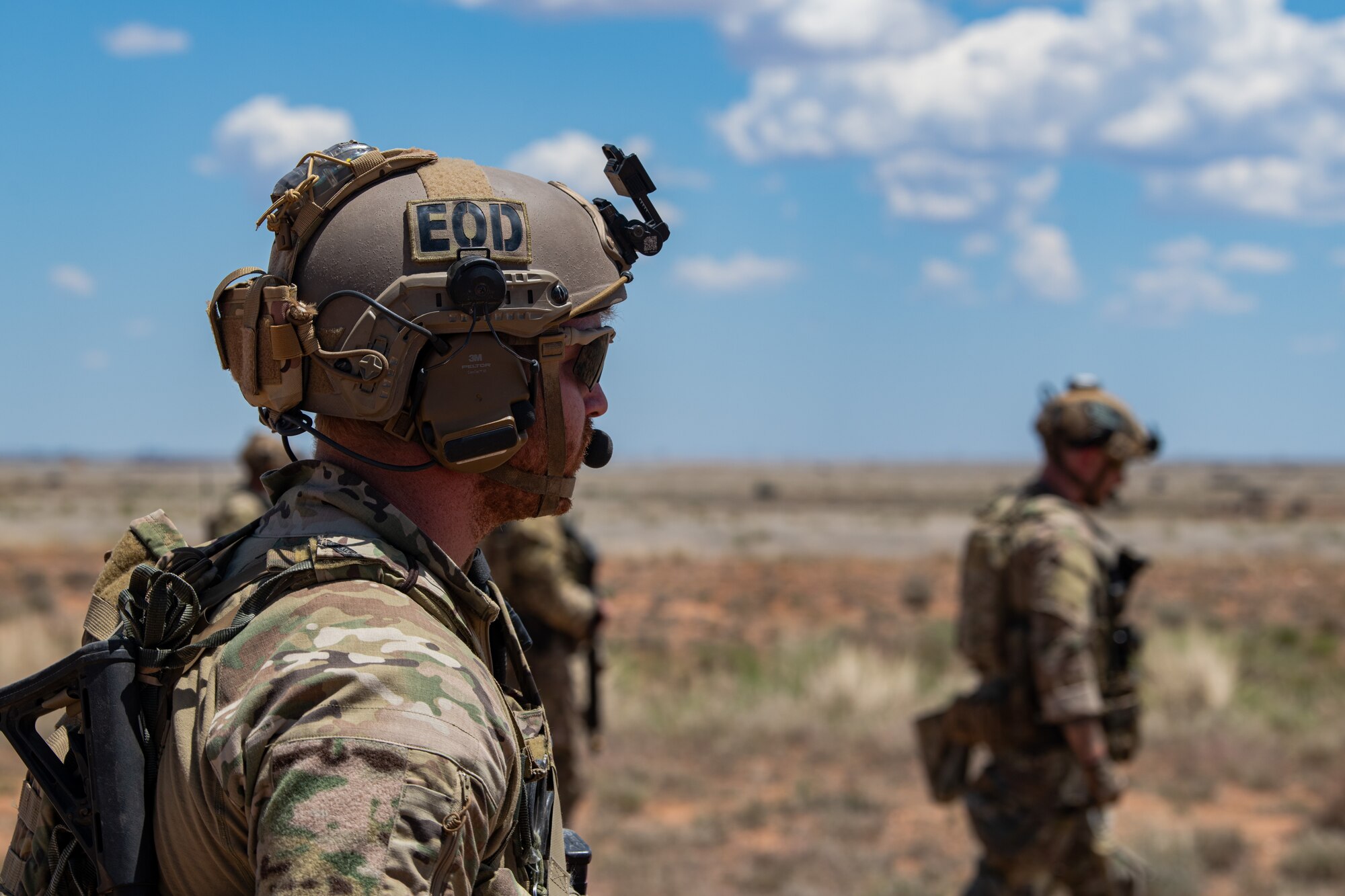 Several men in military uniform and military equipment stand looking out at a plain.