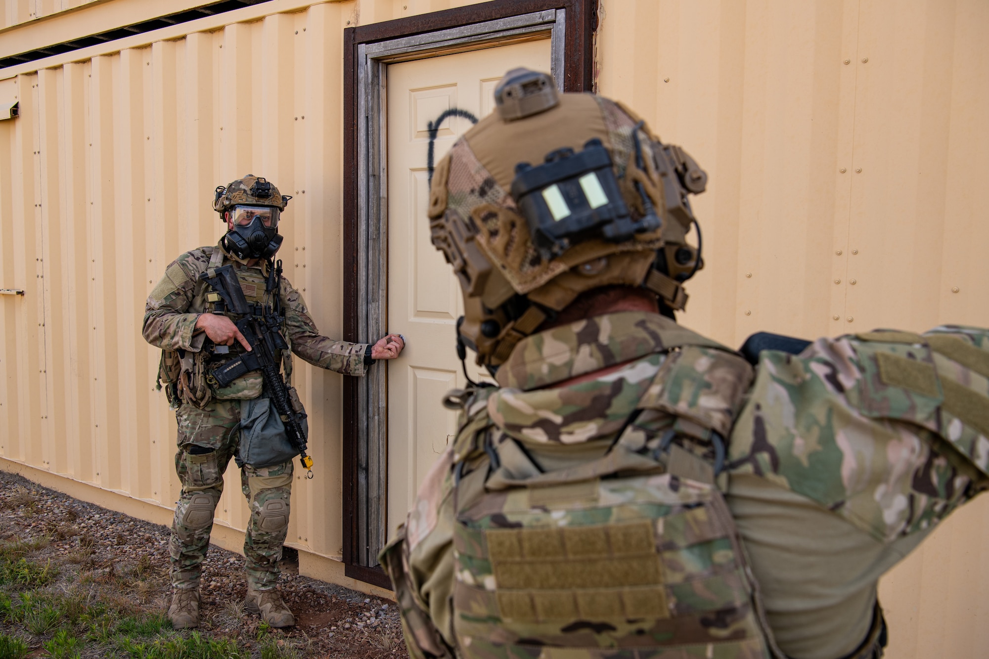 Two men in military uniform with rifles and military equipment prepare to enter a building.