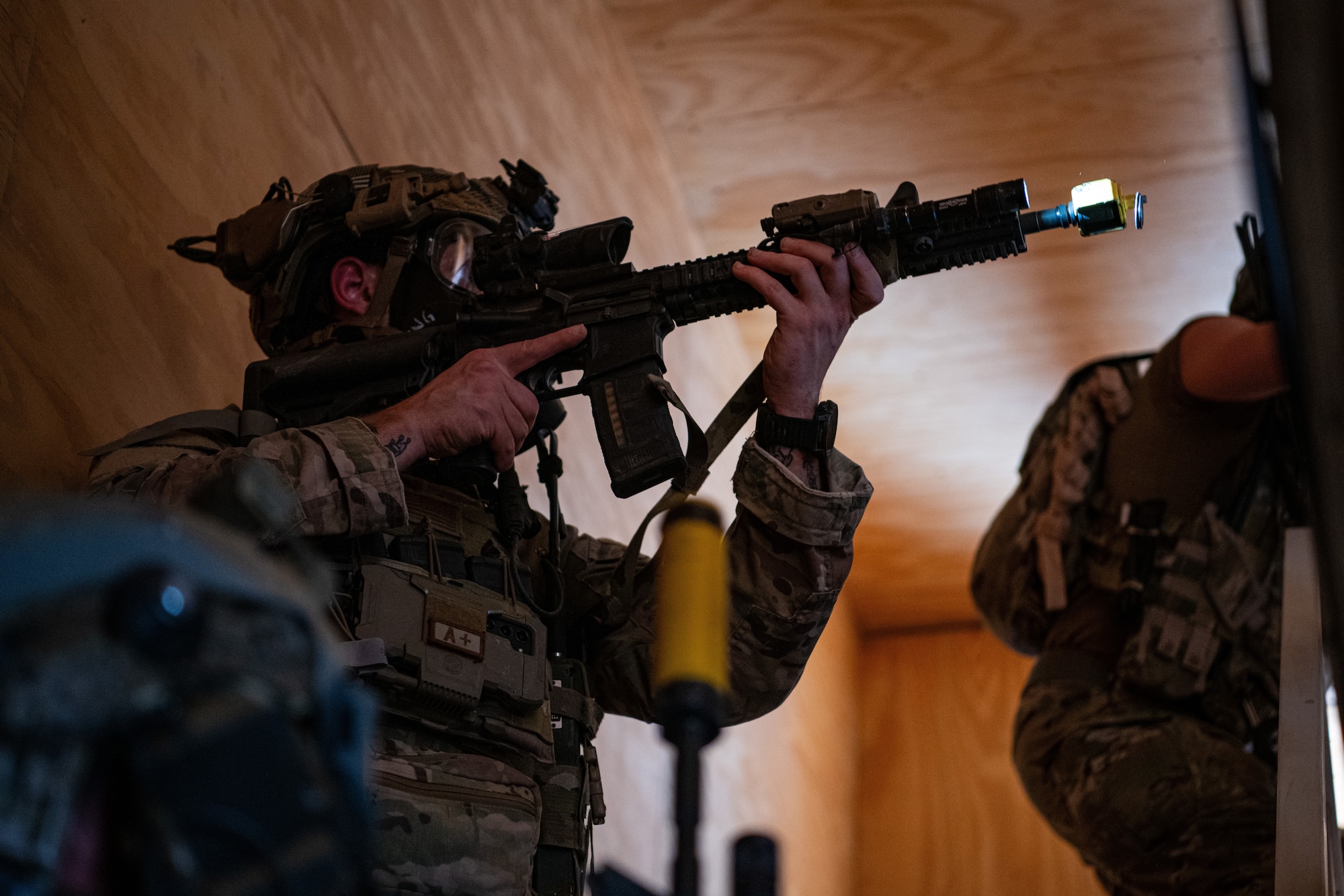 A man in military uniform points a rifle as he ascends a staircase inside a building.