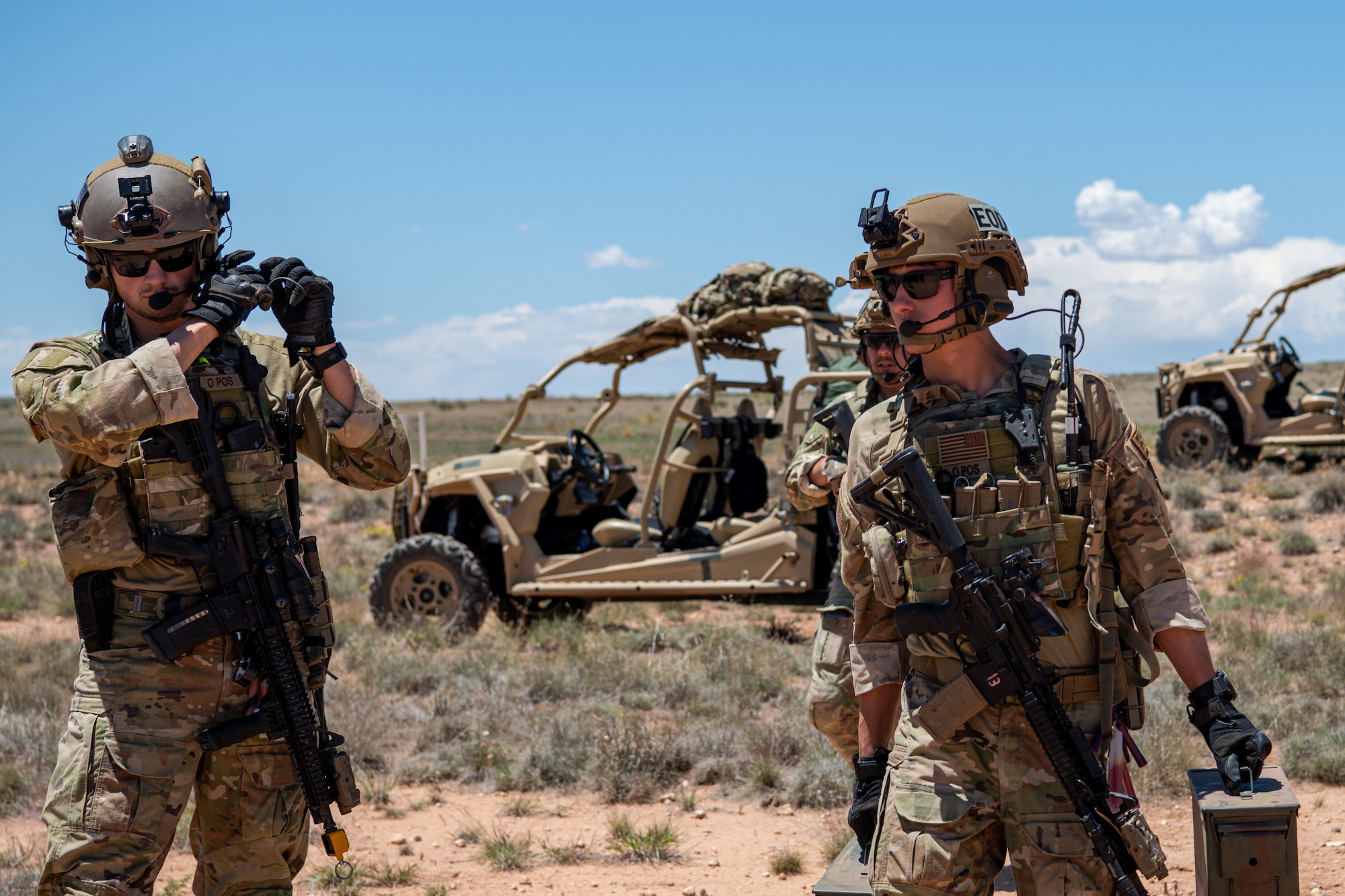 Two men in military uniform and military equipment walk on a plain in front of multiple all-terrain vehicles.
