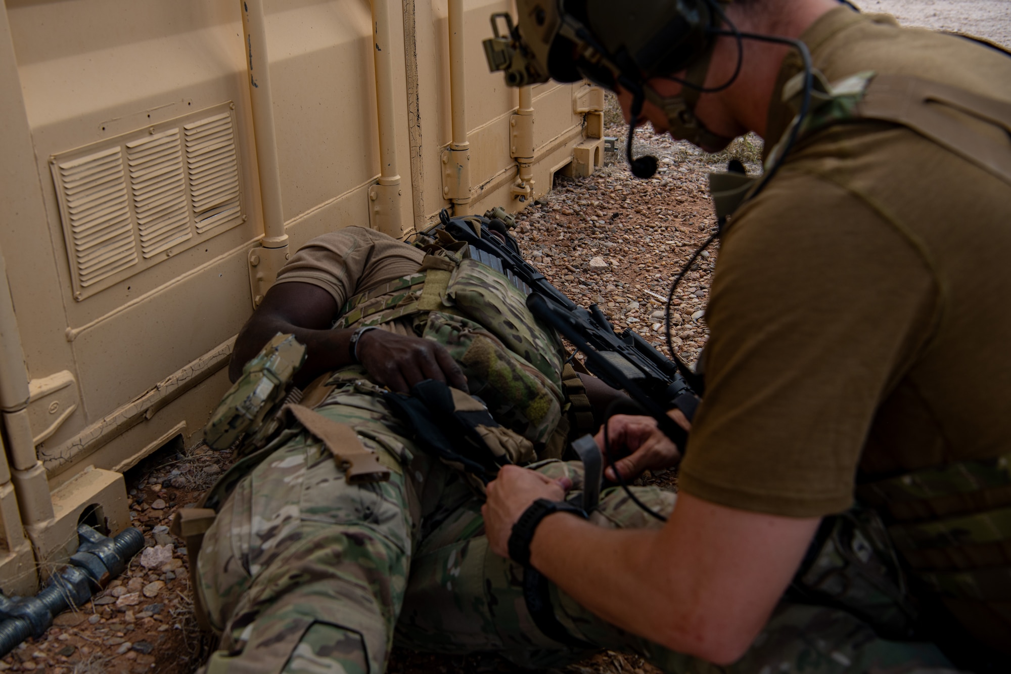 A man in military uniform provides medical aid to another man in military uniform laying on the ground.