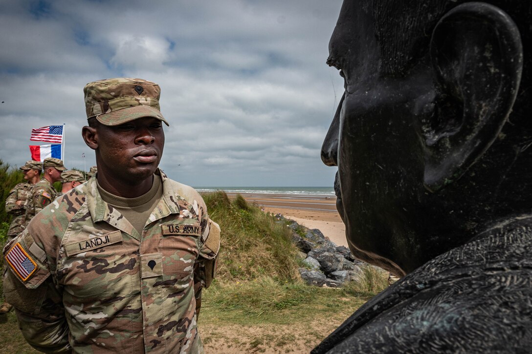 A soldier looks at a stone bust likeness of a veteran positioned on a bluff overlooking the sea.