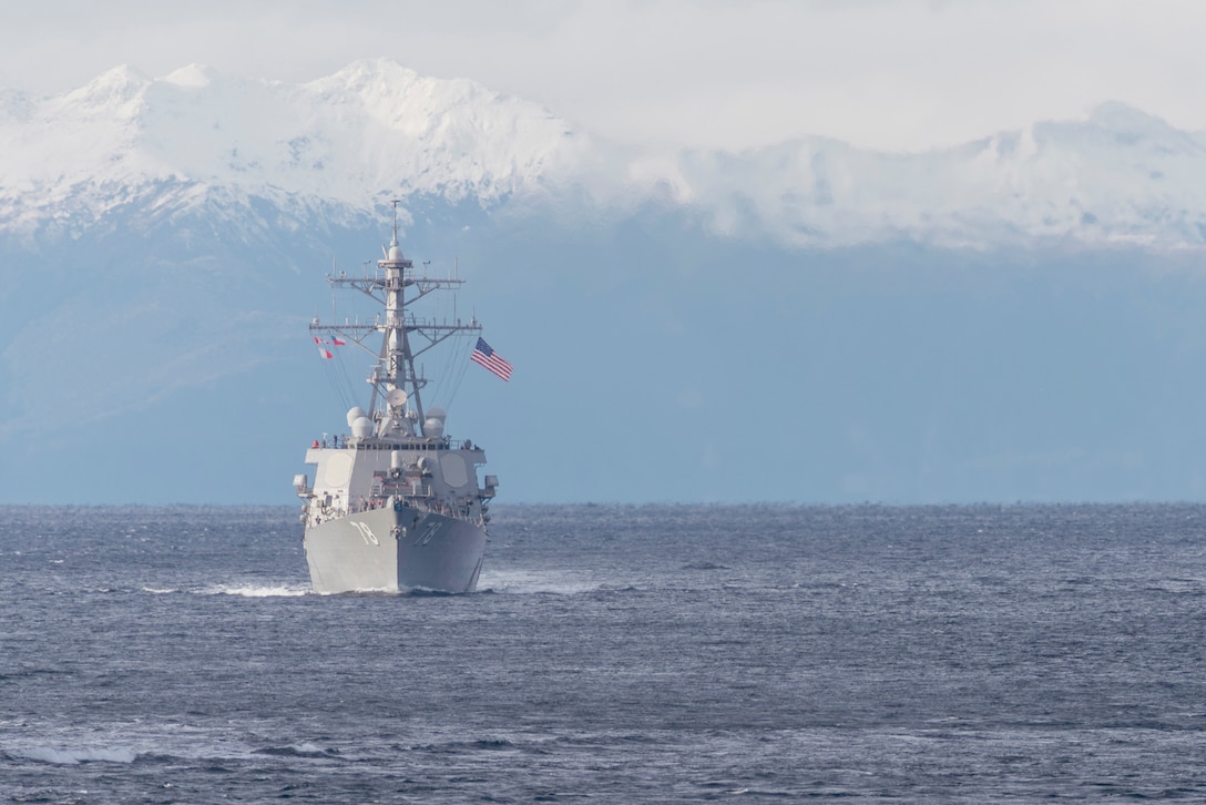 A Navy ship sails in open water with snow-capped mountains in the background.