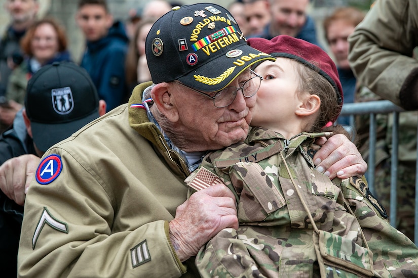 A child kisses the cheek of a seated veteran wearing a “World War II veteran” cap. The veteran is holding the child’s shoulders.