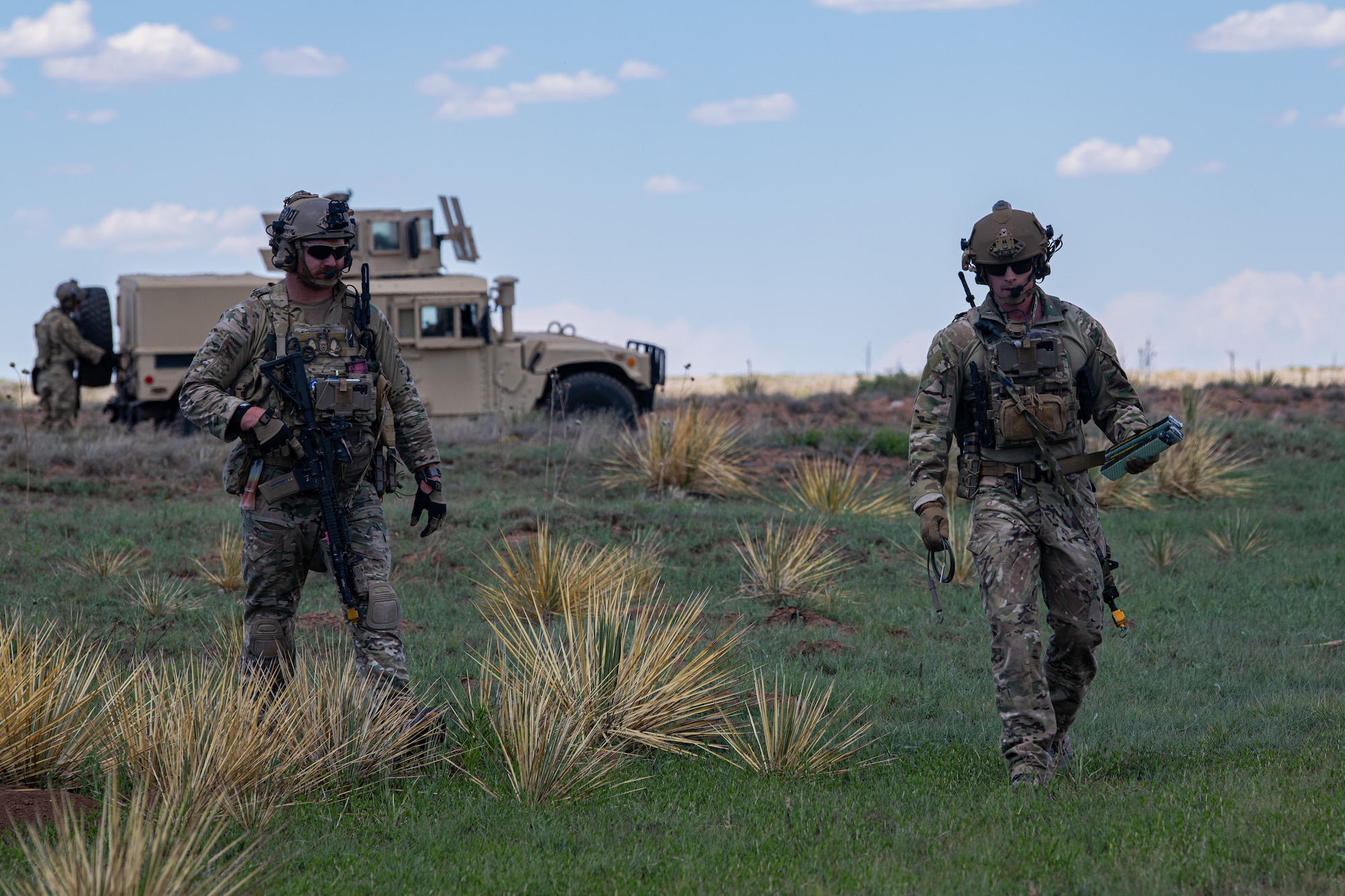 Two men in military uniform walk on a grass plain in front of a military vehicle.