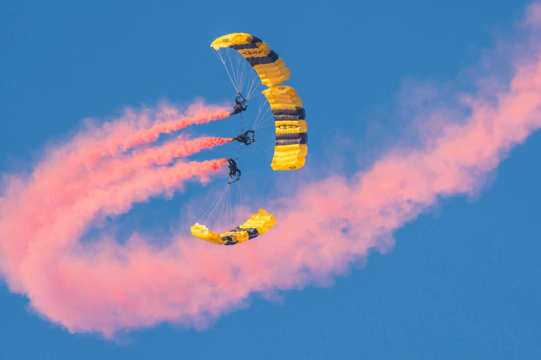 Three soldiers with parachutes perform maneuvers leaving pink streaks in the sky.