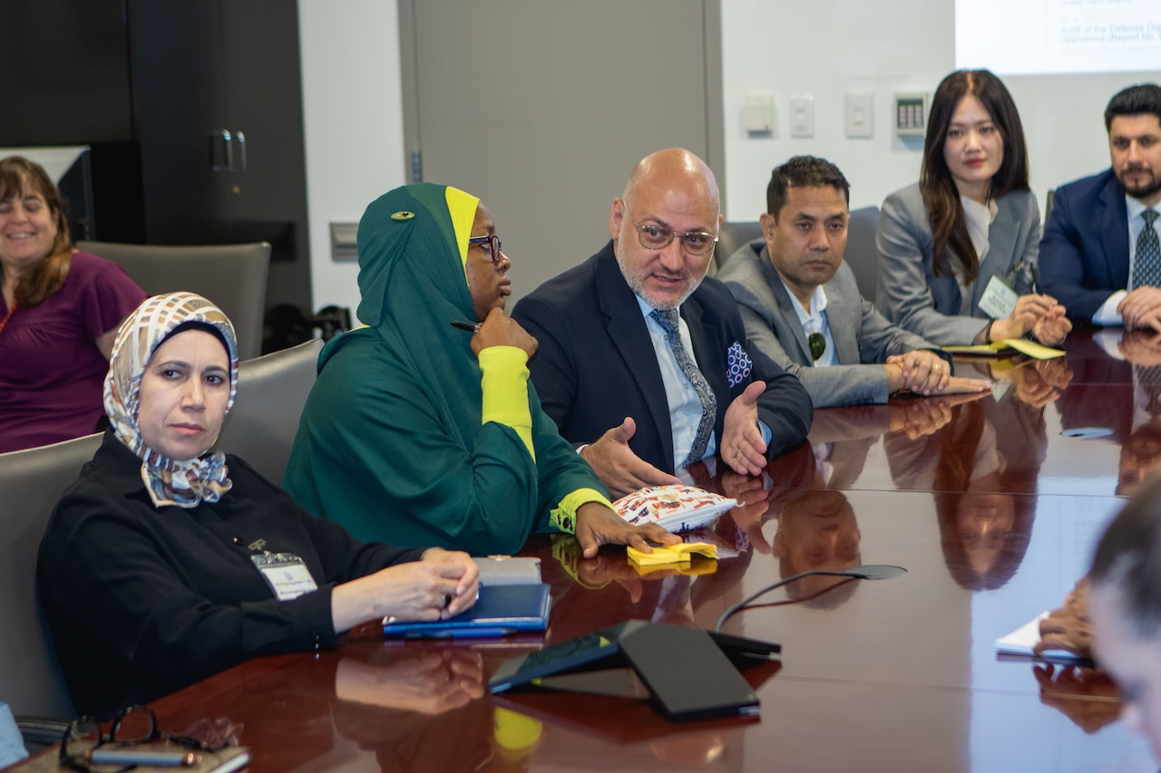 A man in civilian attire gestures with his hands while seated at a conference table next to other men and women in civilian attire.