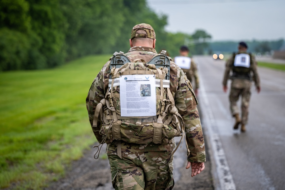 Back image of military member marching with a photo and bio page attached to his rucksack