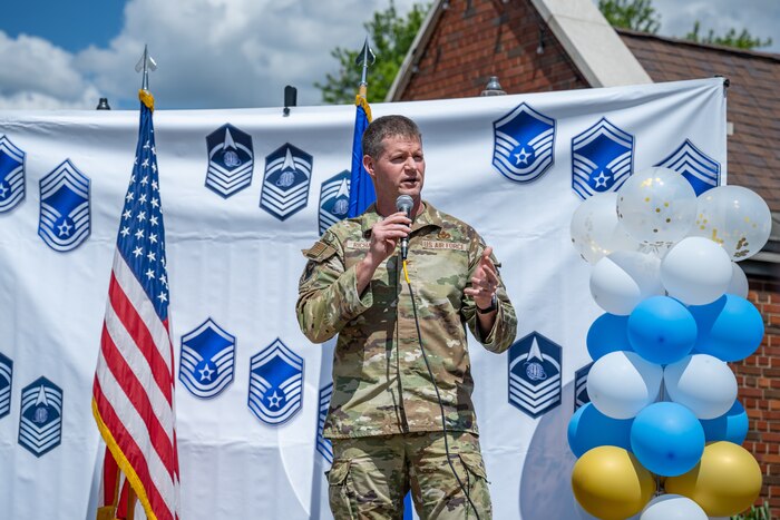 Col Richards gives a speech in front of flags, balloons, and a background with military ranks displayed