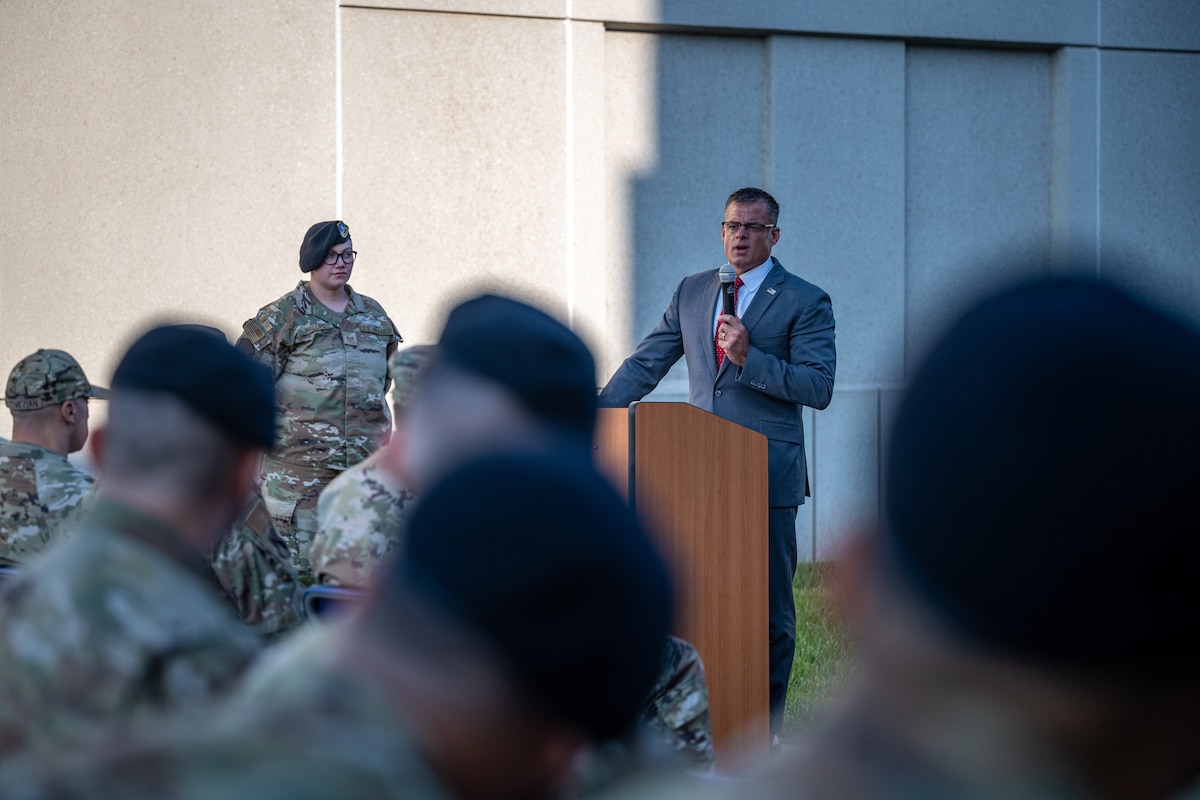 Andy Wilson stands behind a podium giving a speech in a courtyard outdoors with military members sit in the foreground