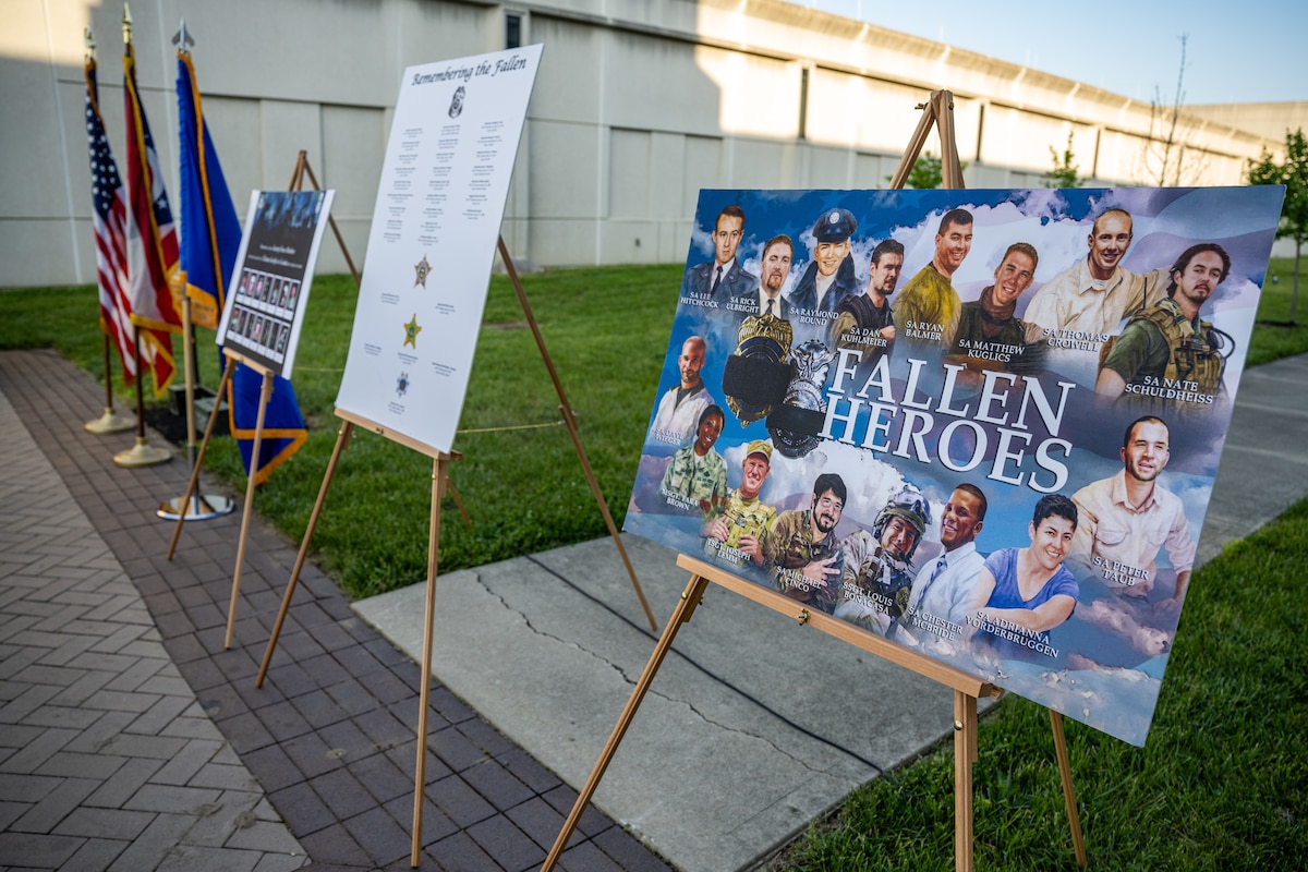 Three signs with photos, illustrations, and names of fallen law enforcement heroes on stands with flags in the background