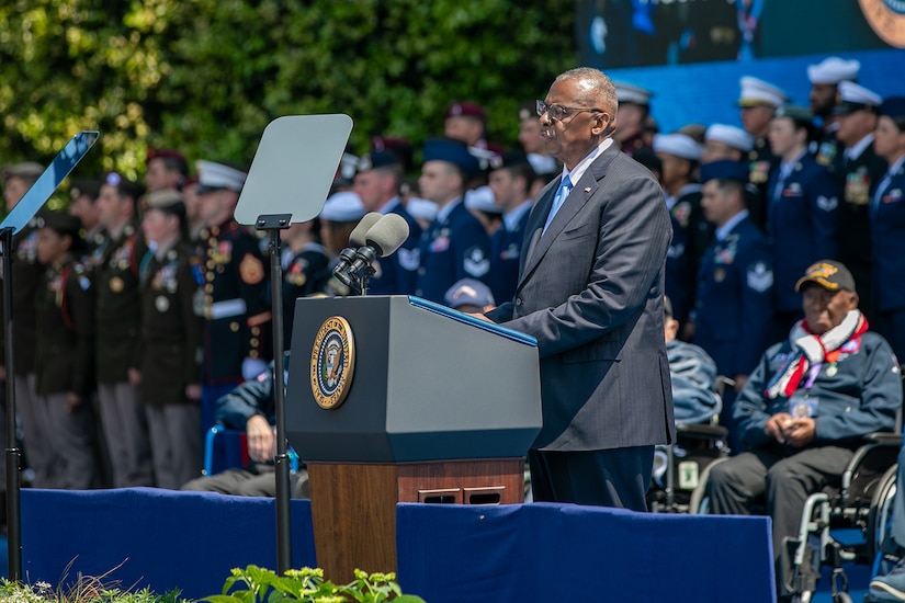 Secretary of Defense Lloyd J. Austin III stands and speaks at a lectern with military and veterans standing and seated behind.