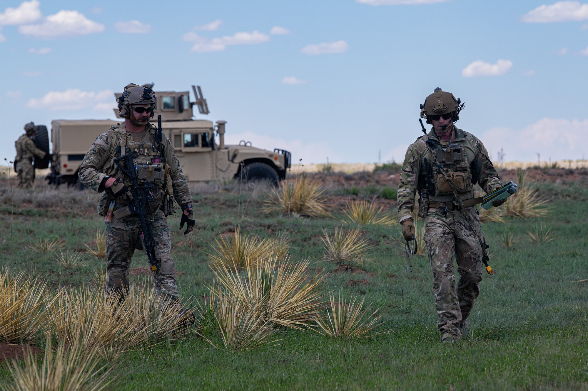 Two men in military uniform walk on a grass plain in front of a military vehicle.