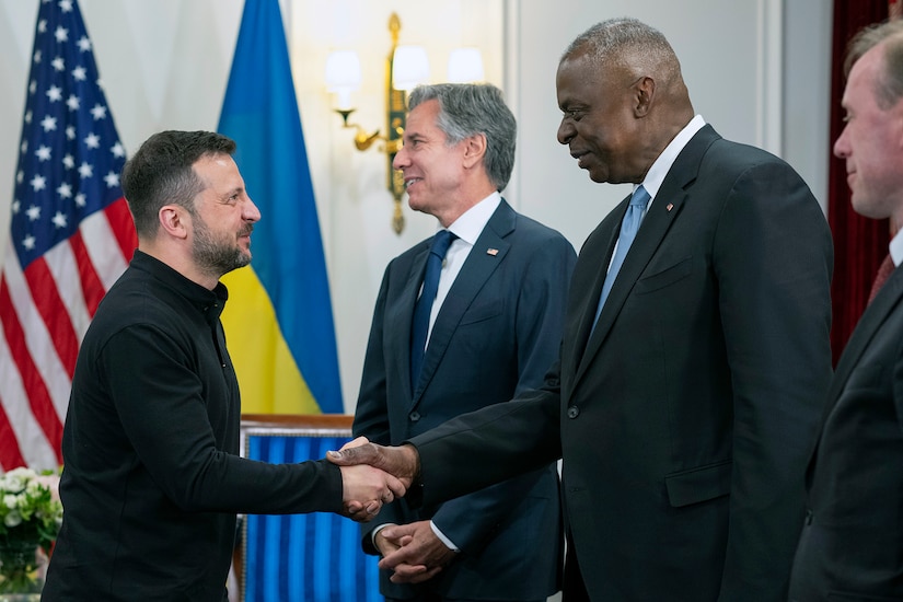 Four people shake hands with flags behind them.