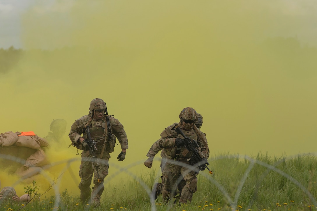 Soldiers carrying weapons run through a field surrounded by yellow clouds of smoke with barbed wire in the foreground.