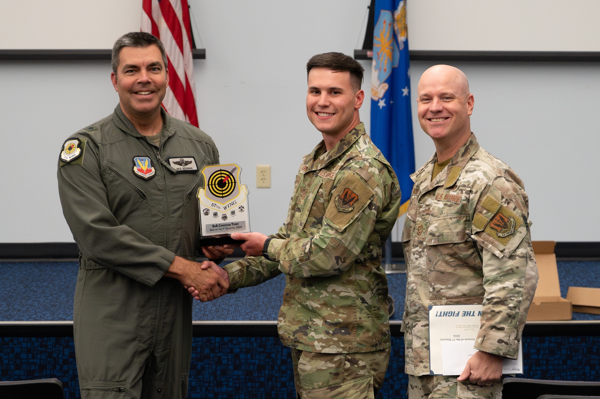 Two men shake hands and hold an award while a third man stands on the side.