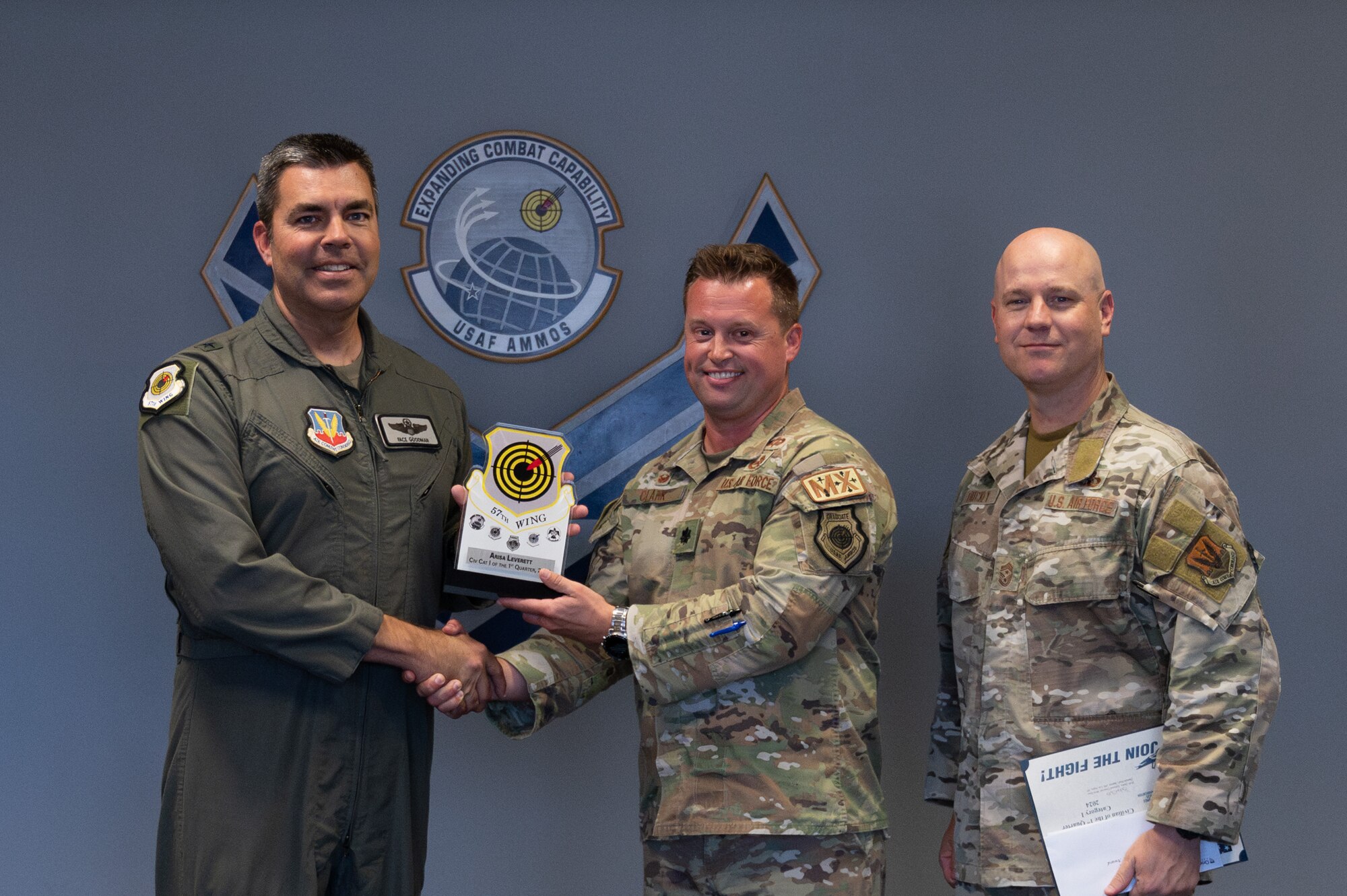 Two men shake hands and hold an award while a third man stands on the side.