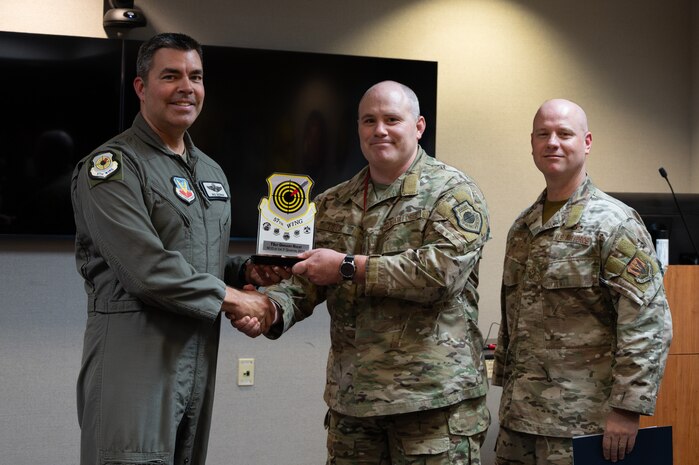 Two men shake hands and hold an award while a third man stands on the side.