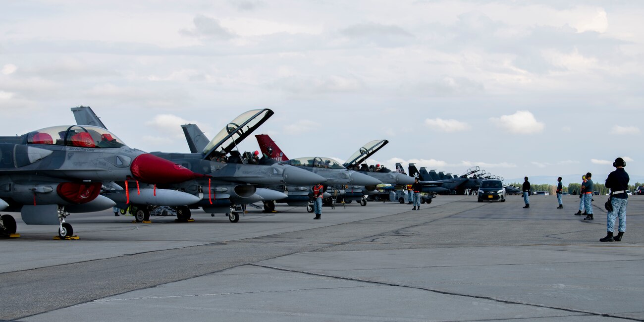4 maintainers from the Republic of Singapore Air Force prepare stand in front of 4 F-16s preparing for takeoff.